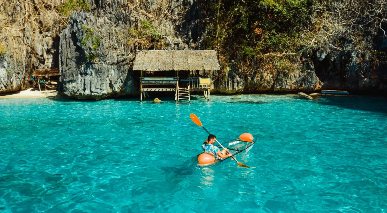 Kayaker paddling through a crystal-clear turquoise lagoon beside a stilted hut and limestone cliffs in Coron