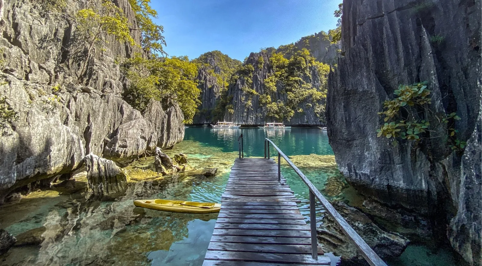 Wooden boardwalk leading through a narrow gap in limestone cliffs into a pristine lagoon with a yellow kayak
