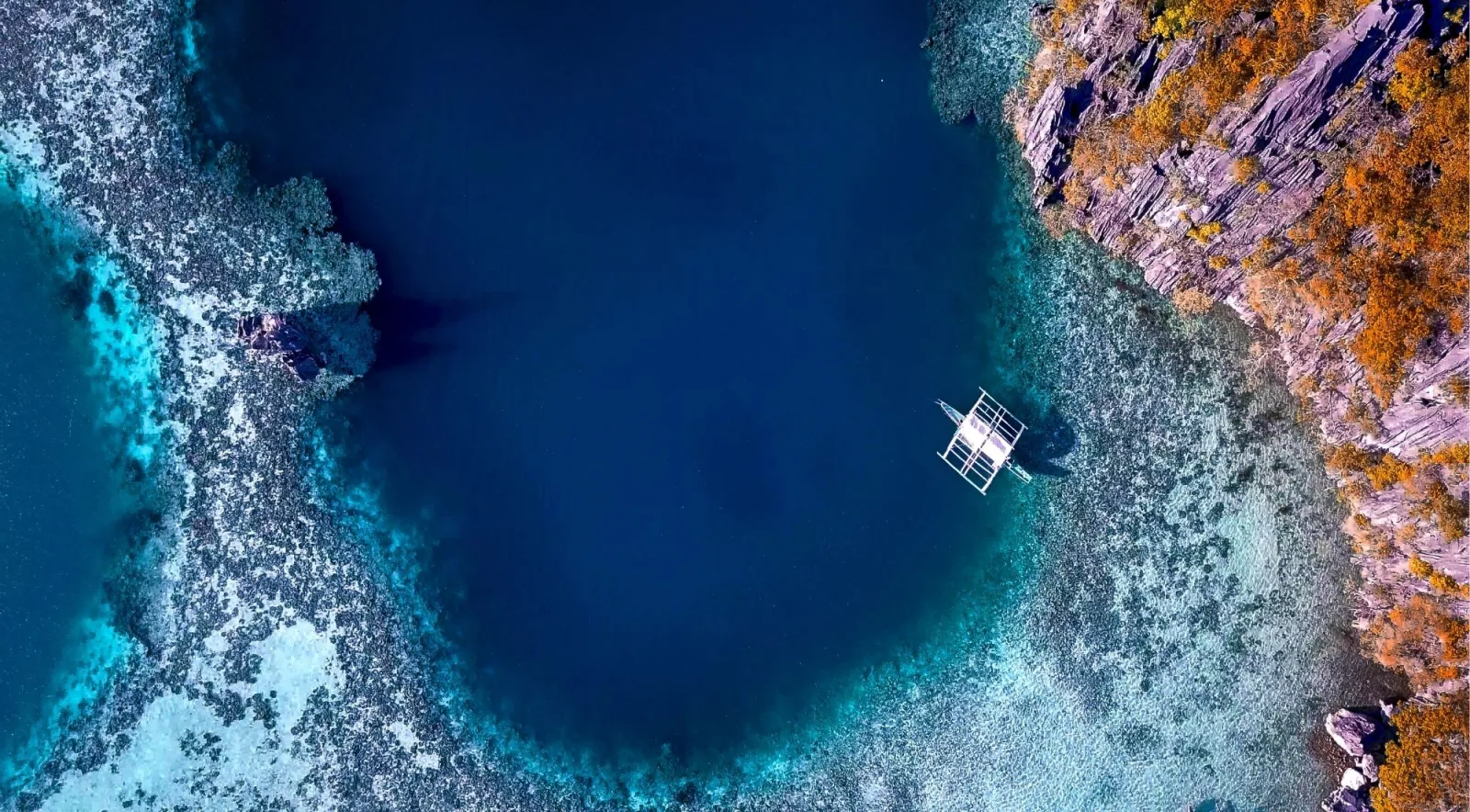 Top-down aerial of a deep blue volcanic crater lagoon surrounded by rust-coloured limestone rocks in Coron